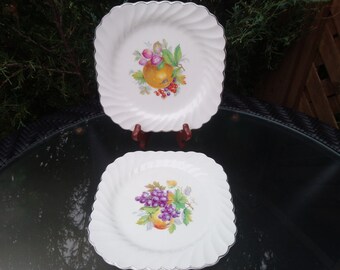 Puede incluir: Dos platos cuadrados de cerámica blanca con borde plateado. Cada plato presenta un diseño de fruta diferente. Un plato tiene una manzana amarilla, uvas y bayas rojas. El otro plato tiene un racimo de uvas moradas y una pera amarilla.