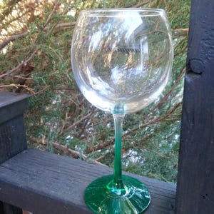 May include: A clear wine glass with a green stem and base. The glass is sitting on a wooden surface with a natural background of green foliage. The glass is empty and reflects the surrounding environment.