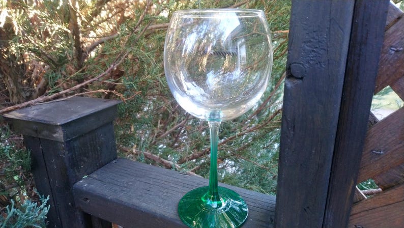 May include: A clear wine glass with a green stem and base. The glass is sitting on a wooden surface with a natural background of green foliage. The glass is empty and reflects the surrounding environment.
