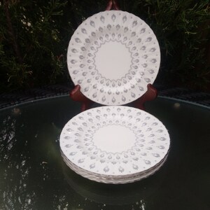 May include: A stack of white ceramic plates with a gray floral pattern. One plate is displayed on a wooden stand. The plates have a circular design with a central medallion and a repeating pattern around the edge. The plates are on a glass table.