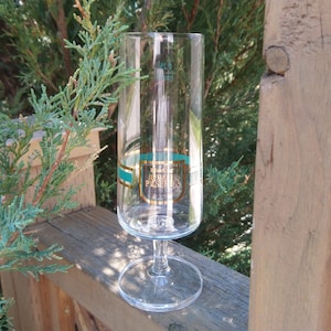 May include: A clear glass champagne flute with a gold and teal band around the middle. The band has the text "La Grande Passion" in gold lettering.