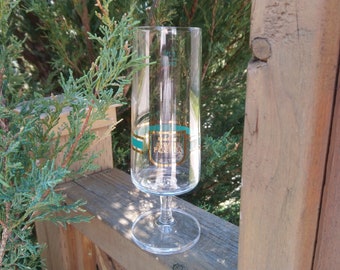 Op de afbeelding: Een heldere glazen champagnefluit met een gouden en turquoise band rond het midden. De band heeft de tekst "La Grande Passion" in gouden letters.