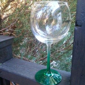 May include: A large, clear wine glass with a green stem and base. The glass has the word "Tang" printed on the side. The glass is sitting on a wooden surface with a natural background.