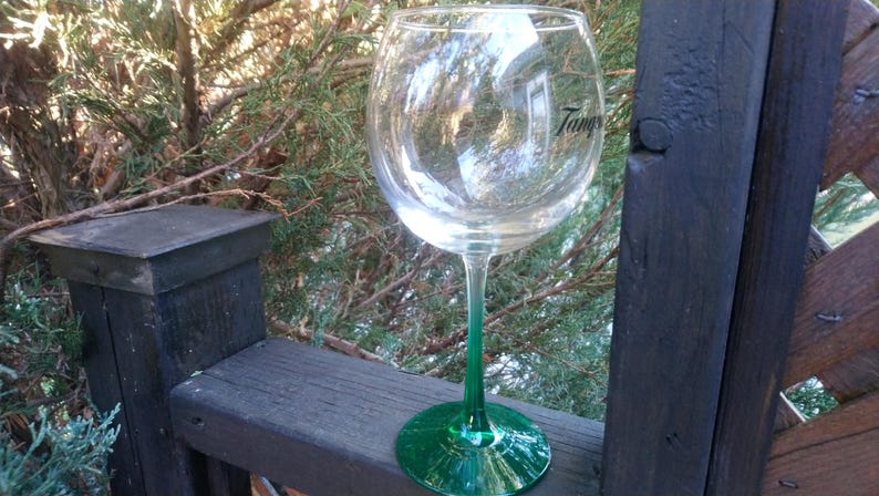 May include: A large, clear wine glass with a green stem and base. The glass has the word "Tang" printed on the side. The glass is sitting on a wooden surface with a natural background.