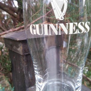 Puede incluir: Un vaso de pinta de vidrio transparente con la palabra "GUINNESS" impresa en blanco en el lateral. El vaso tiene un dise&ntilde;o de arpa en la parte superior.