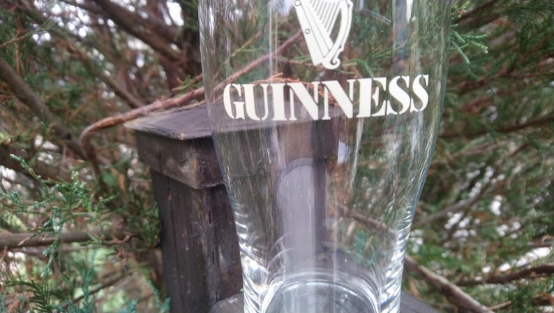 Puede incluir: Un vaso de pinta de vidrio transparente con la palabra "GUINNESS" impresa en blanco en el lateral. El vaso tiene un dise&ntilde;o de arpa en la parte superior.