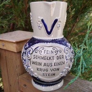 Puede incluir: Una jarra de cer&aacute;mica blanca y azul con un dise&ntilde;o decorativo y el texto "Fein schmeckt der Wein aus einem Krug von Stein".