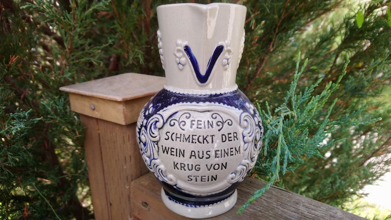 Puede incluir: Una jarra de cer&aacute;mica blanca y azul con un dise&ntilde;o decorativo y el texto "Fein schmeckt der Wein aus einem Krug von Stein".