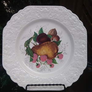 May include: White ceramic square plate with an embossed design. The centre of the plate features a painted design of a pear, plum, and nuts with pink raspberries.