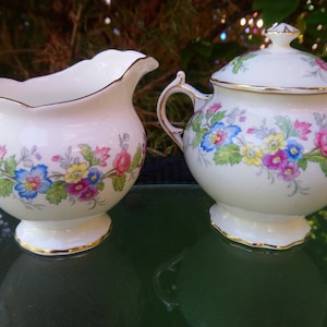 May include: A white creamer and sugar set with a floral design in shades of pink, blue, yellow, and green. The set has gold trim around the edges.