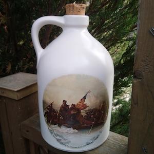 May include: White ceramic jug with a cork stopper and a painted image of a historical scene depicting a group of people in a boat with a flag. The image is titled "Crossing the Delaware River 1776".
