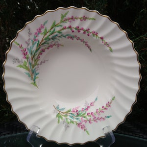 May include: A white ceramic bowl with a gold rim and a floral design featuring pink and green flowers. The bowl has a scalloped edge and is ideal for serving soup, salad, or dessert.