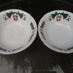Peut inclure: Deux bols en céramique blanche avec un bord doré. Les bols présentent un motif écossais vert, rouge et or avec un blason au centre. Les bols sont sur une surface sombre.
