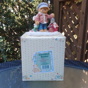 以下が含まれることがあります： 帽子、コート、ジーンズを着てランタンを持っているテディベアの装飾的な置物。クマは小さな家と別のクマがいる雪のベースに立っています。置物は「Cherished Teddies」と書かれた箱の上にあります。