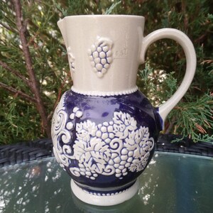 Puede incluir: Una jarra de cer&aacute;mica blanca con un dise&ntilde;o de uvas azul y blanco. La jarra tiene un asa y un pico. Est&aacute; decorada con uvas y hojas en relieve.