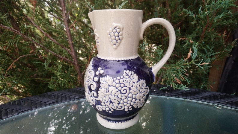 Puede incluir: Una jarra de cer&aacute;mica blanca con un dise&ntilde;o de uvas azul y blanco. La jarra tiene un asa y un pico. Est&aacute; decorada con uvas y hojas en relieve.