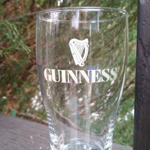 Puede incluir: Un vaso de pinta de vidrio transparente con la palabra "GUINNESS" impresa en blanco en la parte delantera. Tambi&eacute;n se imprime un logotipo de arpa en el vaso.