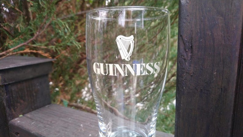 Puede incluir: Un vaso de pinta de vidrio transparente con la palabra "GUINNESS" impresa en blanco en la parte delantera. Tambi&eacute;n se imprime un logotipo de arpa en el vaso.