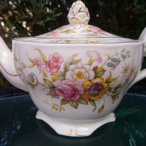 Puede incluir: Una tetera blanca con un diseño floral. La tetera tiene una tapa con un pomo en forma de capullo de flor. La tetera tiene un borde dorado alrededor del borde y la base.