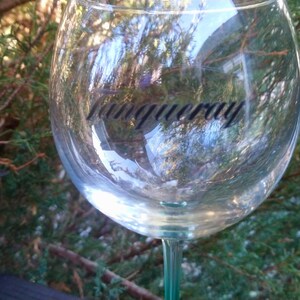 May include: A clear, stemmed gin glass with the word "Tanqueray" printed in black. The glass is round and reflects the surrounding greenery. The stem is a light green color.
