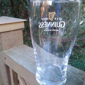 May include: A clear glass pint glass with the Guinness logo printed on the side. The logo is in gold and white and reads "Guinness" with the text "Brewed in Dublin" and "1778" below. The glass is sitting on a wooden surface.