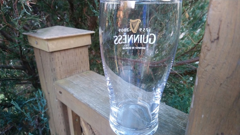 May include: A clear glass pint glass with the Guinness logo printed on the side. The logo is in gold and white and reads "Guinness" with the text "Brewed in Dublin" and "1778" below. The glass is sitting on a wooden surface.