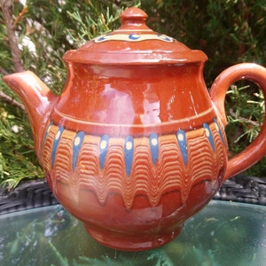 Op de afbeelding: Een bruine theepot van keramiek met een deksel, versierd met blauwe en gele geometrische patronen. De theepot heeft een rond lichaam en een gebogen handvat.