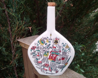 May include: White ceramic bottle with a cork stopper. The bottle is shaped like a diamond and features a colorful illustration of the city of Munich, Germany. The text "München" is printed on the bottom of the bottle.