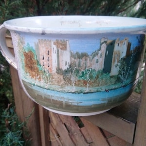 May include: A white ceramic bowl with a hand-painted scene of a castle and a river. The bowl has a blue and green glaze and a brown handle.