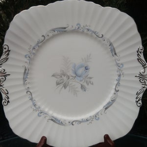 Puede incluir: Plato de cena de cerámica blanca con borde festoneado y borde plateado. El centro del plato presenta un diseño floral azul y gris con una sola rosa.