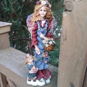 May include: A porcelain figurine of two angels, one larger than the other. The larger angel is wearing a blue denim dress with a red shirt underneath and a floral wreath. She is holding a basket of flowers. The smaller angel is wearing a blue dress with a red shirt underneath and a floral wreath. Both angels are standing on a cloud.