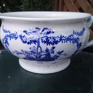 Puede incluir: Un orinal de cerámica blanca con un diseño floral azul y blanco. El orinal tiene una asa y una base ancha.