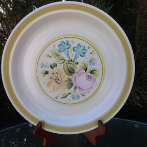 May include: A white ceramic dinner plate with a yellow rim and a floral design featuring pink, yellow, and blue flowers. The flowers are painted in a vintage style.