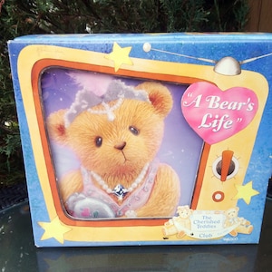 Puede incluir: Una caja decorativa con una ilustración de un oso de peluche. El oso lleva un collar de perlas y una tiara, con el texto "A Bear's Life" en un gráfico en forma de corazón. La caja tiene un fondo azul con estrellas y un marco amarillo.
