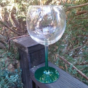 May include: A large, clear wine glass with a green stem and base. The glass has the word "Maurry" printed on the side. The glass is sitting on a wooden surface, with green foliage in the background.