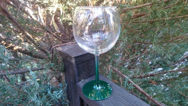 May include: A large, clear wine glass with a green stem and base. The glass has the word "Maurry" printed on the side. The glass is sitting on a wooden surface, with green foliage in the background.