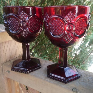 Puede incluir: Dos copas de vidrio rojo rubí con un patrón de diamante y un diseño en relieve. Las copas están sobre una superficie de madera.