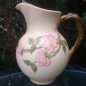 Puede incluir: Una jarra de cerámica blanca con asa marrón y un diseño floral con rosas rosas y hojas verdes.