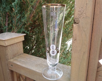 Op de afbeelding: Een hoog, helder glazen bierglas met een gouden rand en een zwart-wit logo. Het glas heeft een ronde basis en een hoog, slank lichaam.