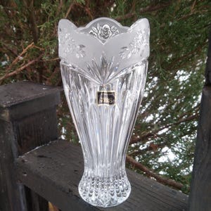Peut inclure: Un vase en cristal transparent avec un motif floral givré sur le dessus. Le vase a une forme évasée et une base texturée. Une petite étiquette noire et dorée est visible à l'avant. Le vase mesure environ 25 cm de haut.