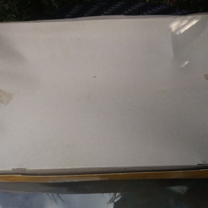 May include: A rectangular cardboard box with a light beige lid, sealed with tan tape. The box is slightly damaged, with a dented corner. The box is sitting on a glass table, with greenery in the background.