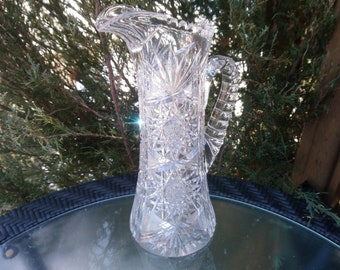 May include: A clear glass pitcher with a cut glass design. The pitcher has a tall, slender body and a handle.