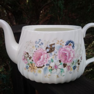 May include: White teapot with a floral design featuring pink, blue, and yellow flowers. The teapot has a handle and a spout.