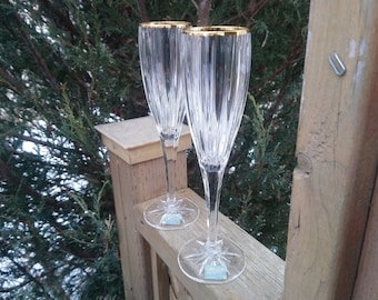Può includere: Due eleganti flûte da champagne con bordi dorati. Le flûte in vetro trasparente hanno un design a coste verticali e basi a forma di stella. Sono ambientate su uno sfondo di fogliame verde e pali di legno.