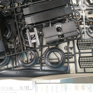 May include: A model kit with various black plastic parts, including tires, body panels, and other components. The parts are still attached to their plastic frames, ready for assembly. The kit is a 1:24 scale model.