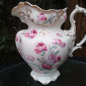 May include: A white porcelain pitcher with a floral design. The pitcher has a gold trim and a handle with a decorative design. The flowers are pink, red, and purple.