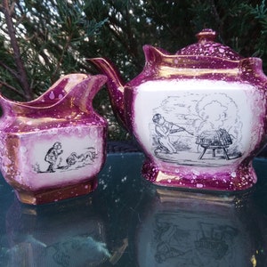 May include: A pink and white teapot and creamer set with black and white illustrations. The teapot has a handle and spout. The creamer has a handle. Both pieces have a splatter pattern.