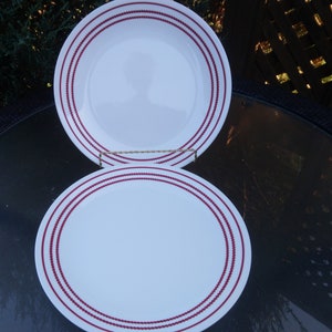 May include: Two white ceramic plates with a red dotted border. The plates are round and are displayed on a glass table. The plates are a classic design for everyday use.