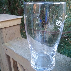 May include: A clear glass pint glass with the word "Guinness" printed on the side. The glass is empty and sitting on a wooden surface.