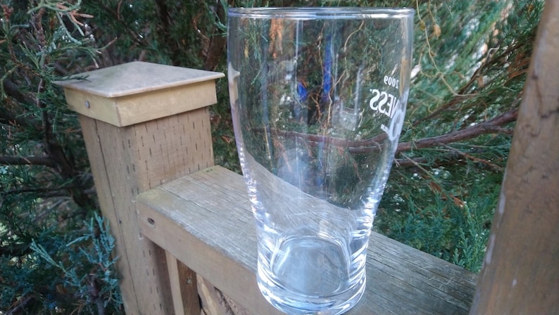 May include: A clear glass pint glass with the word "Guinness" printed on the side. The glass is empty and sitting on a wooden surface.
